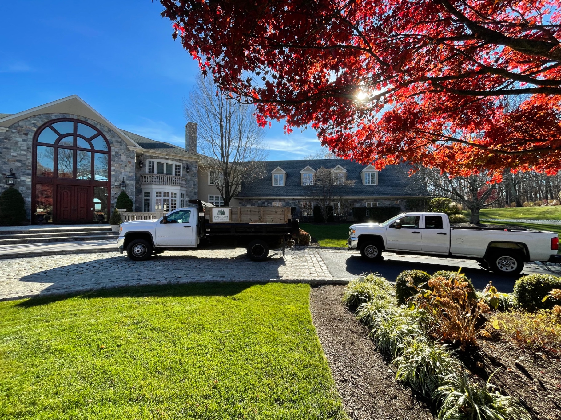 Fortress Landscaping trucks parked on a cobblestone motor court at a stone estate in autumn
