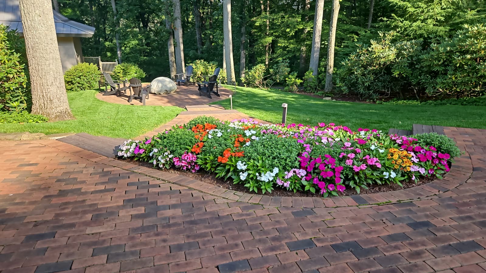 Curved bed of impatiens and zinnias in mixed colors set into a brick paver patio beside a wooded lawn